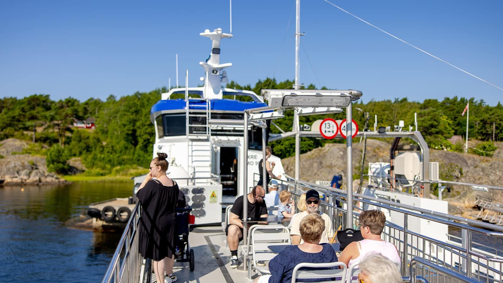 Passasjerer sitter og nyter solen på dekk på Hvalerferga ved kai på Nordre Sandøy, med grønn vegetasjon i bakgrunnen.