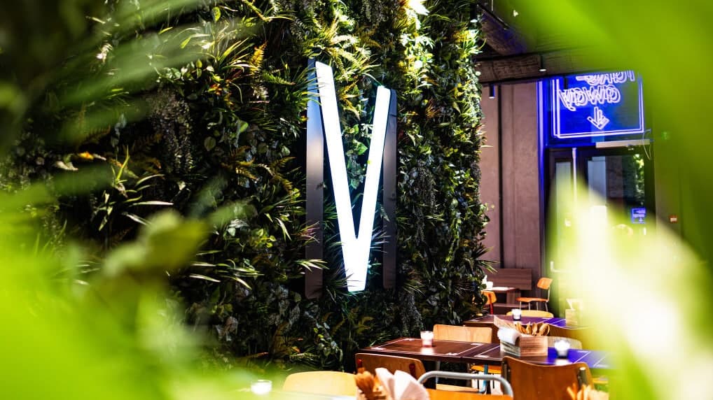 Grønn vegg med lysende M-logo i Matværste, omgitt av bord og stoler i moderne interiør.