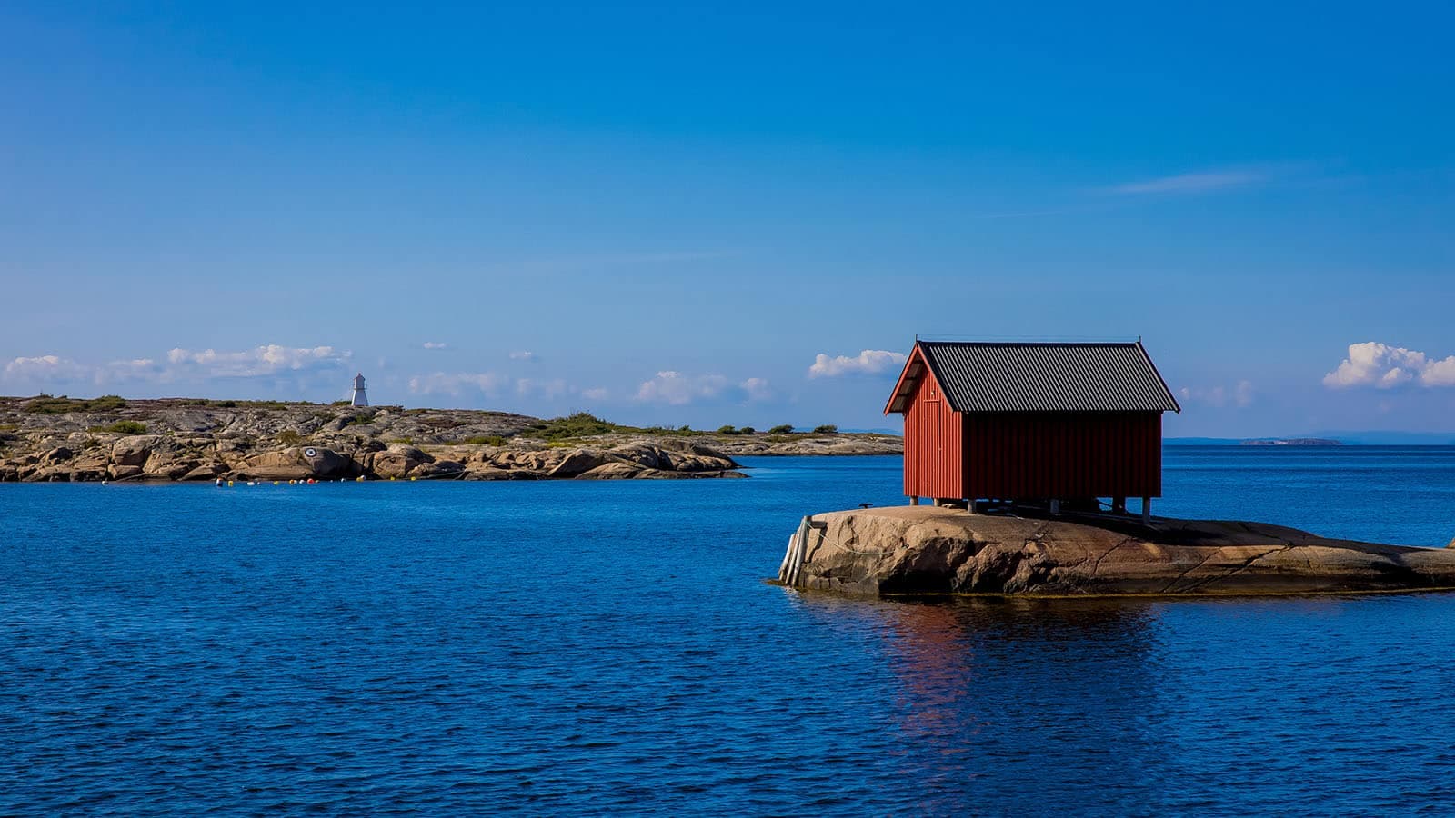 Rød sjøbod på et svaberg i sjøen ved Papperhavn på Vesterøy, Hvaler, med fyrlykt i bakgrunnen og blå himmel over kystlandskapet.