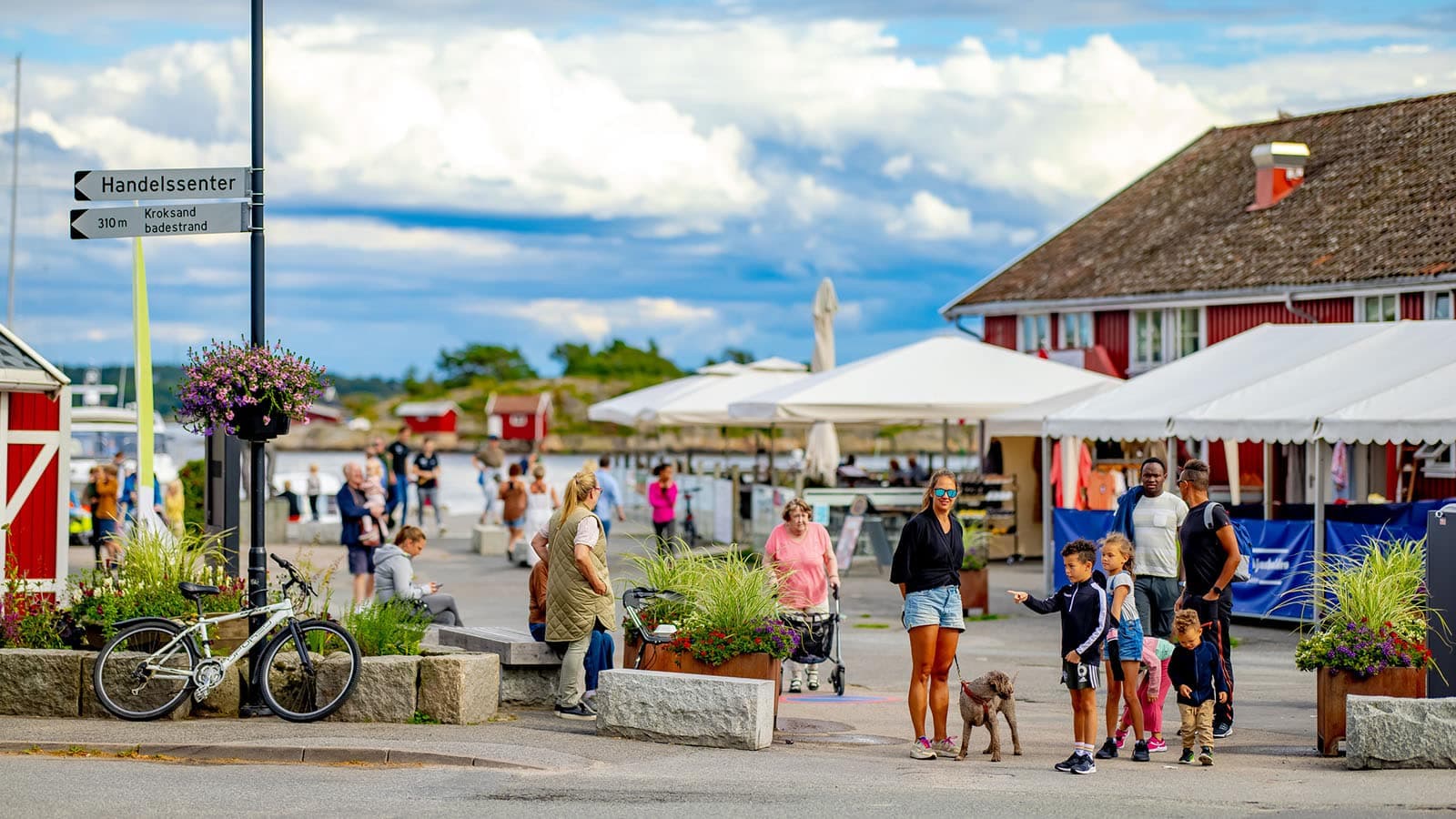 Mennesker rusler mellom boder, butikker og spisesteder på Skjærhalden, med sykkelparkering, blomster og båthavn i bakgrunnen.