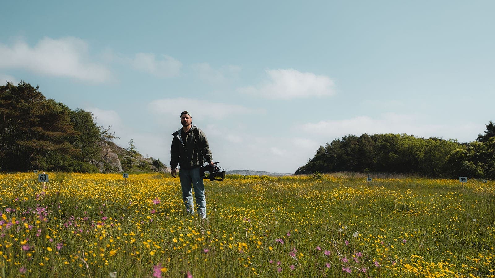 Person med filmkamera i blomstereng på Storesand, Hvaler, omgitt av natur og blå himmel.