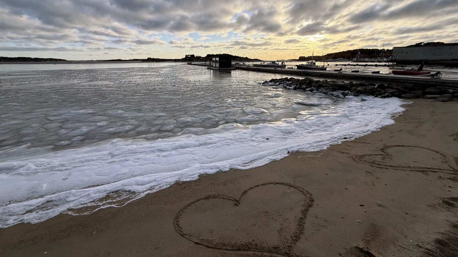 Hjerte tegnet i sanden på vinterstrand på Hvaler, med islagt sjø, brygge og mykt vinterlys i bakgrunnen.