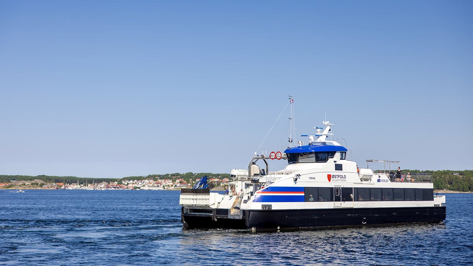 Fergen "Torine" på vannet med passasjerer om bord, blå himmel og Skjærhalden i bakgrunnen.