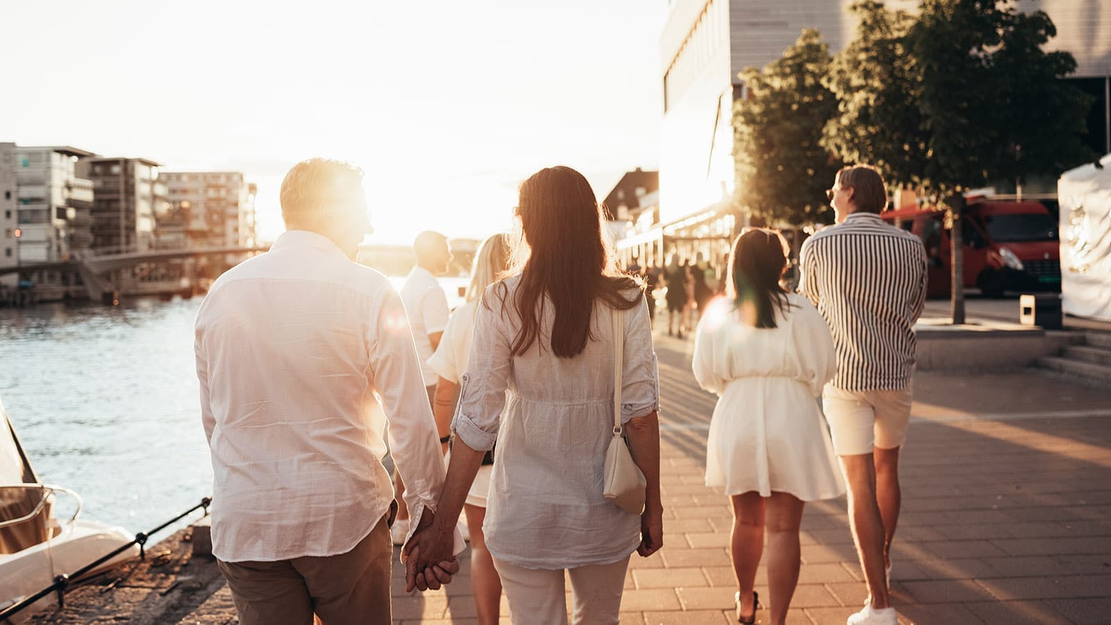 En varm sommerkveld ved brygga i Fredrikstad, der folk går langs elvepromenaden i kveldssol. Bildet viser den livlige stemningen ved vannkanten, omgitt av restauranter, båtliv og moderne bymiljø.