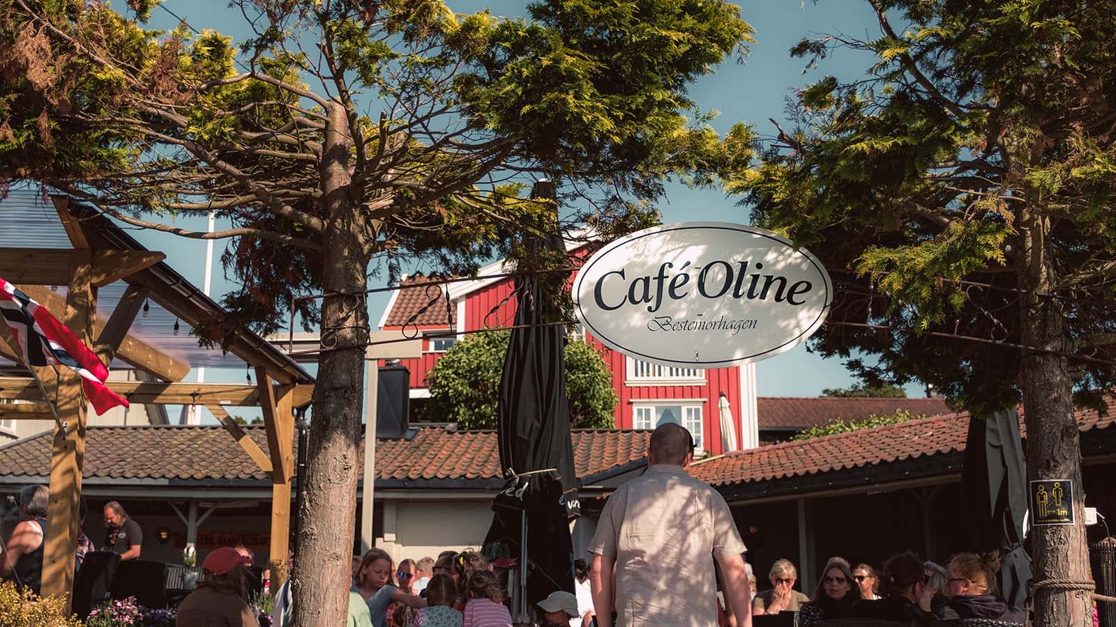 Skilt og uteservering ved Café Oline på Søndre Sandøy, omgitt av trær og folk i sommersol