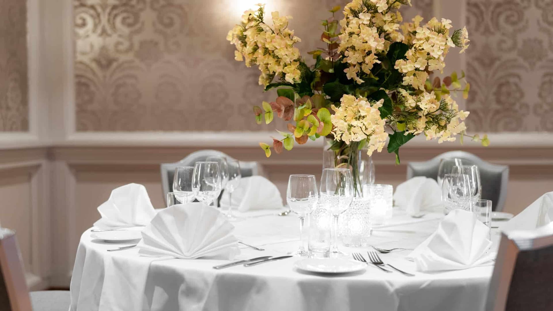 Pent dekket rundt bord med hvit duk, servietter og blomsterdekor i selskapslokale i Fredrikstad.